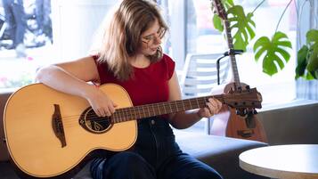 Una persona tocando una guitarra acústica en una habitación con un ventanal grande, una planta y una mesa redonda.