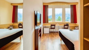 Habitación de hotel con dos camas, un escritorio, un televisor y ventanas grandes con cortinas.