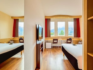 Ein Hotelzimmer mit zwei Betten, einem Schreibtisch, einem Fernseher und großen Fenstern mit Vorhängen.