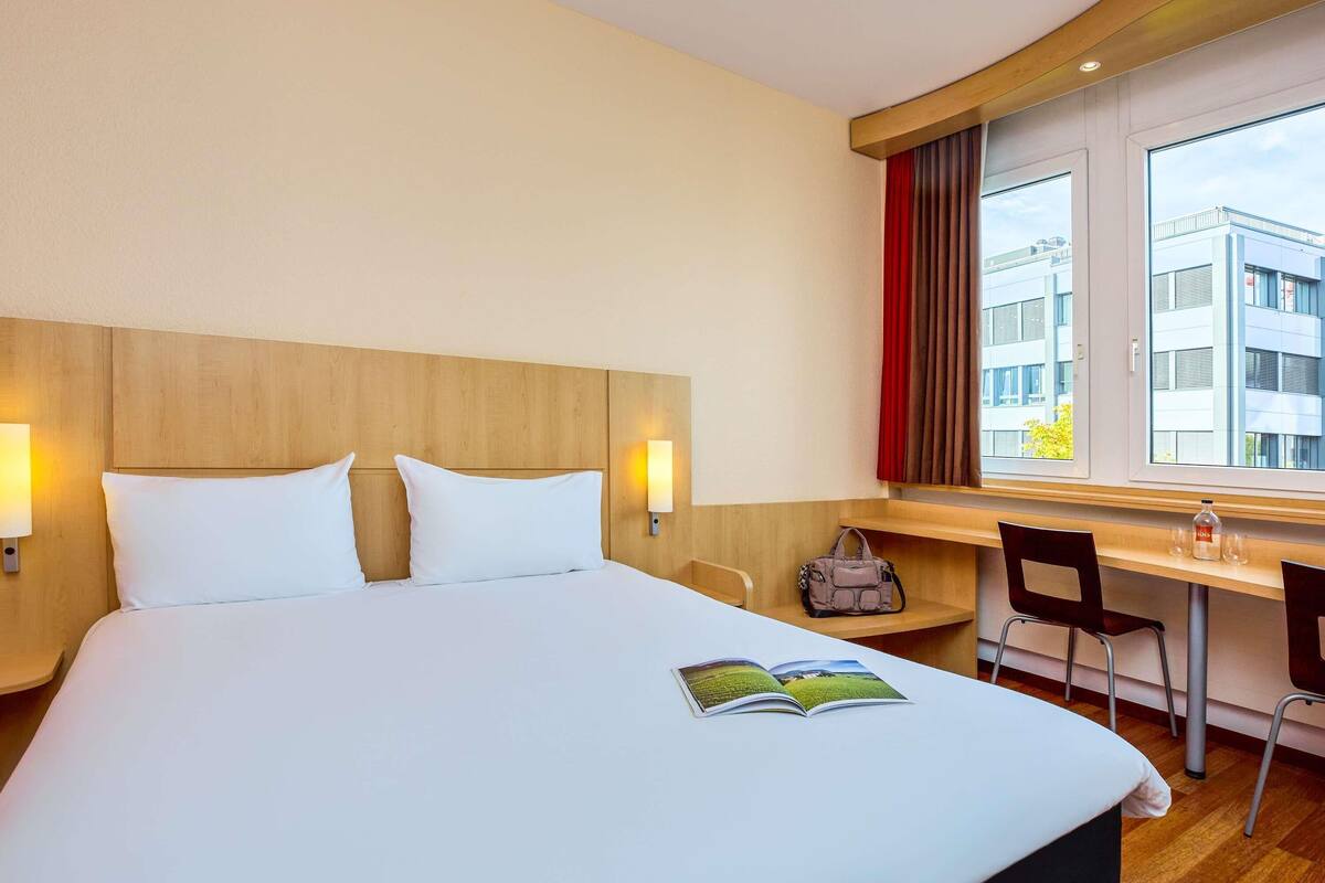 Une chambre d’hôtel avec un grand lit, deux lampes de chevet, un bureau avec une chaise et une fenêtre donnant sur des immeubles.