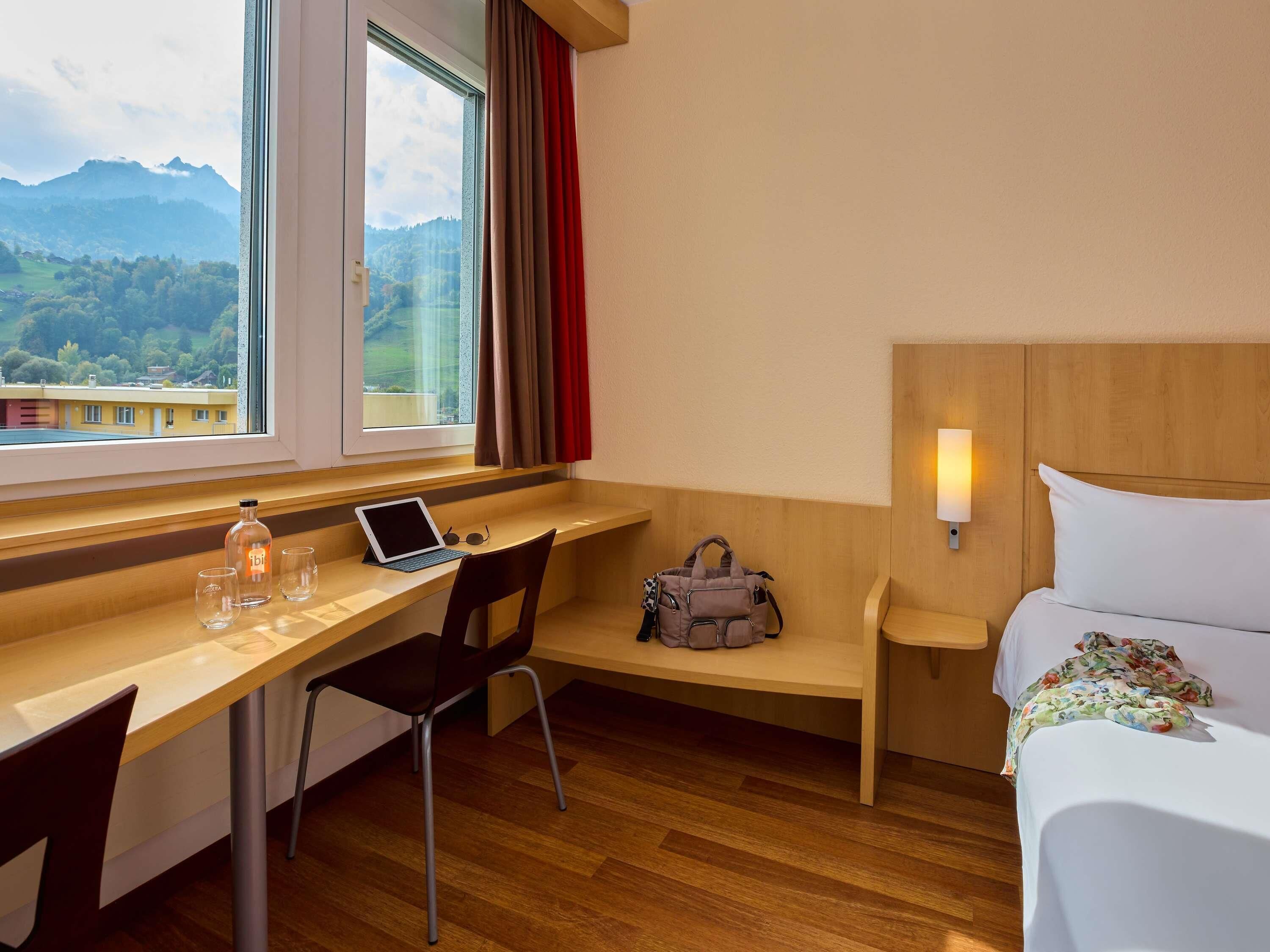 A hotel room with a wooden desk, a laptop, a chair, a bed with a floral bedspread, and a view of mountains through a window.