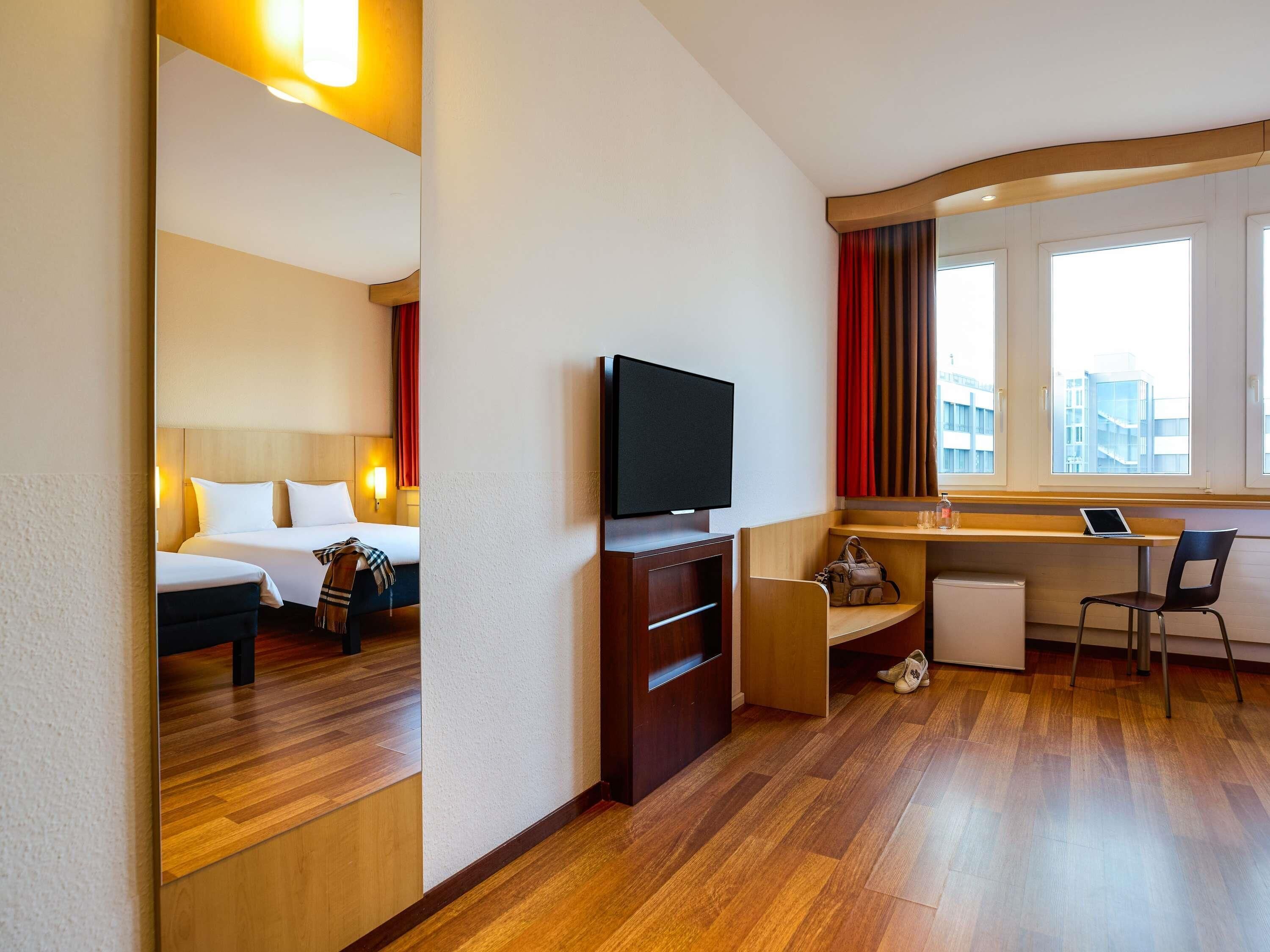 Quarto de hotel com cama, televisão, secretária com um computador portátil e janela com cortinas.