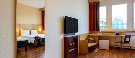A hotel room with a bed, a TV, a desk with a laptop, and a window with curtains.