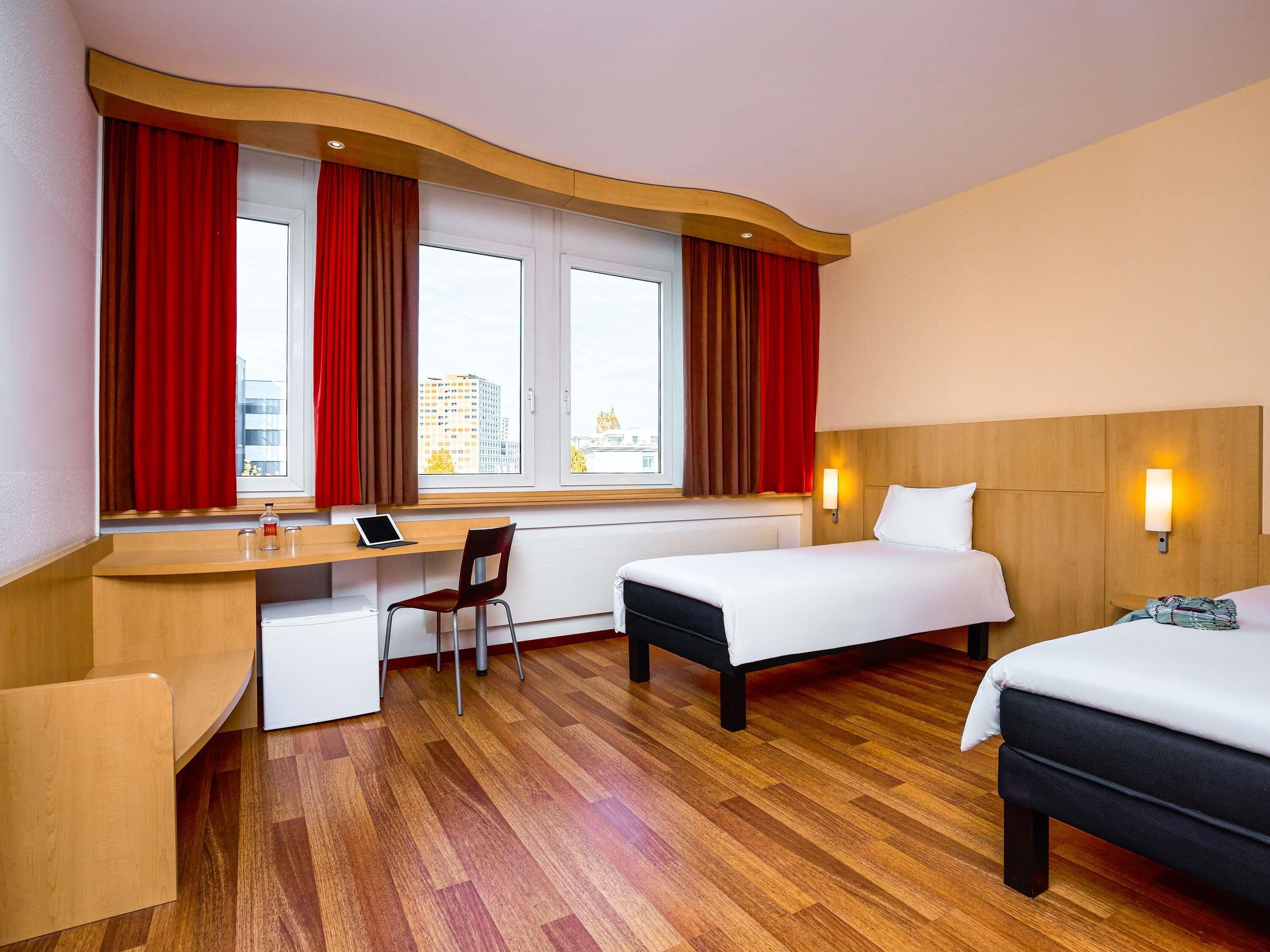 A hotel room with a desk, chair, two beds, and a window with curtains.
