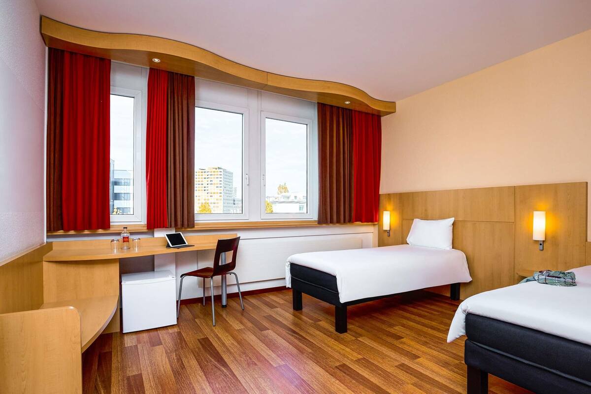 A hotel room with a desk, chair, two beds, and a window with curtains.