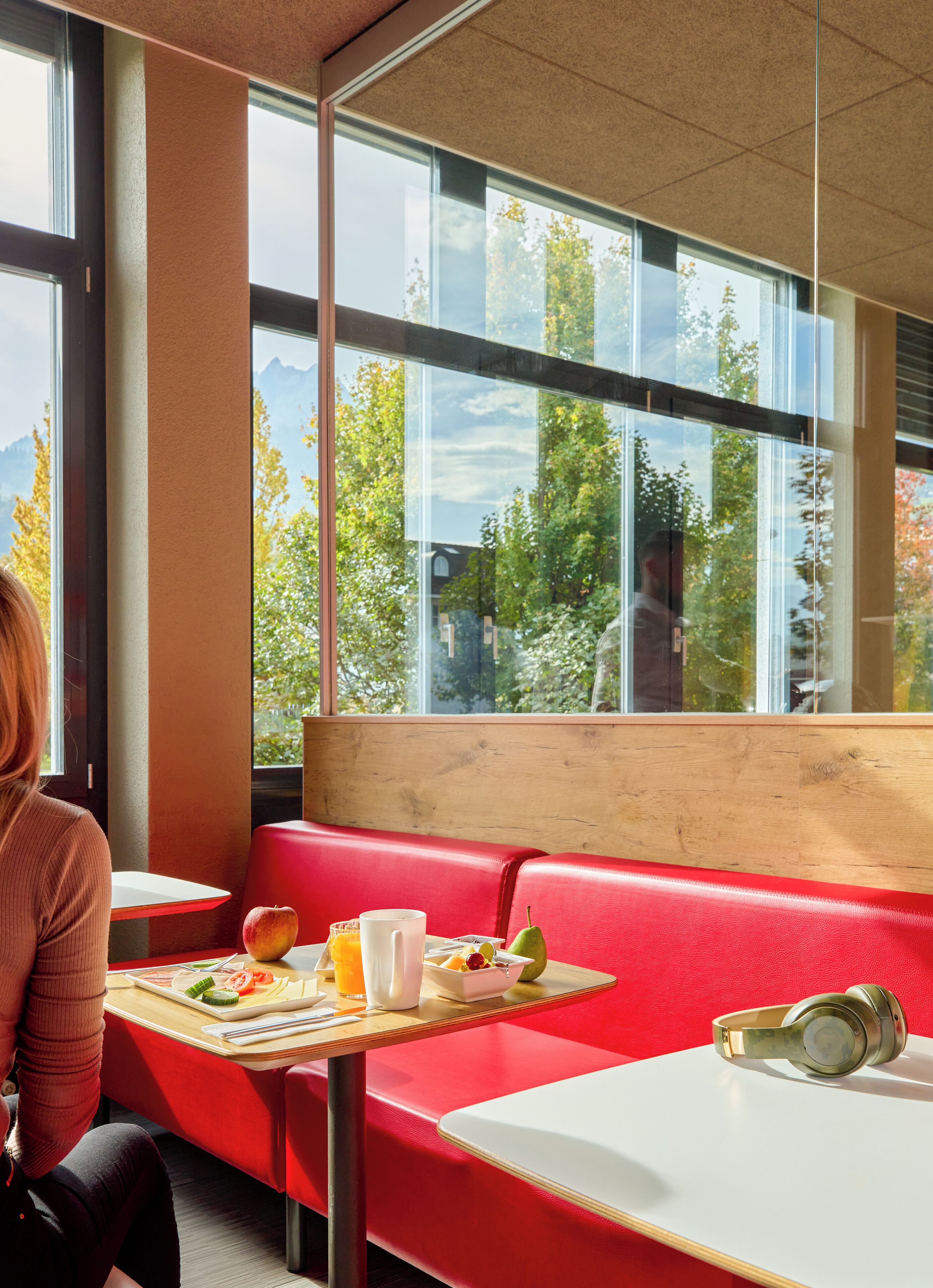 Un interior moderno con un banco y una mesa rojos, una ventana con vistas a árboles y montañas, y una persona sentada a la mesa.