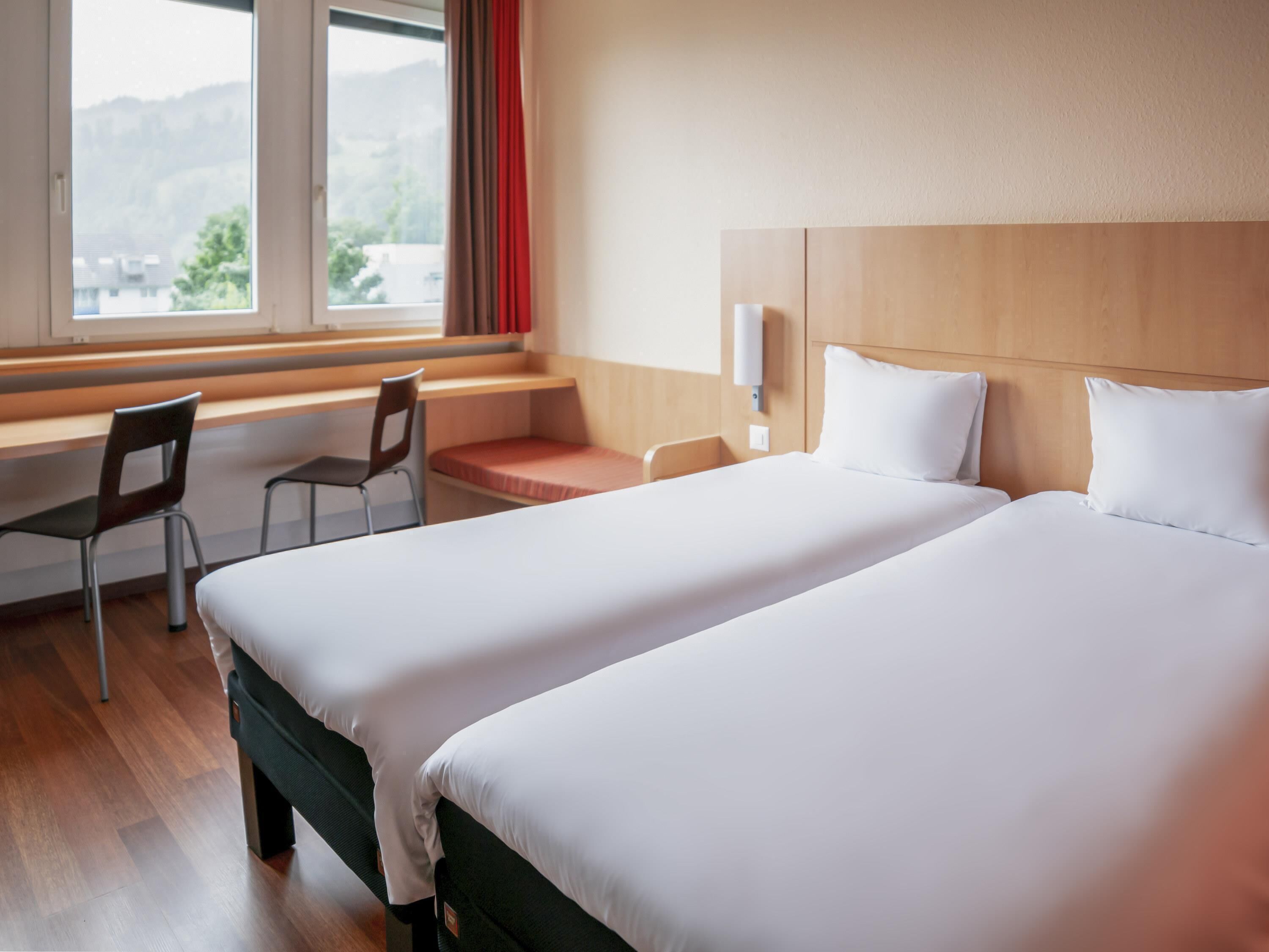 A hotel room with two beds, a desk, a chair, and a window with a view of the outdoors.