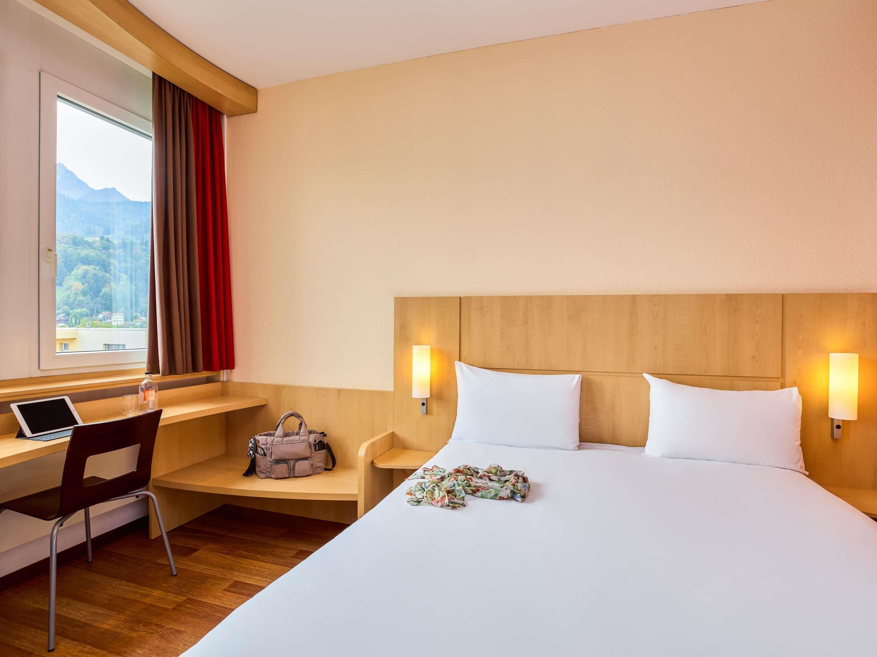 Habitación de hotel con cama, escritorio, silla y vista a las montañas desde la ventana.