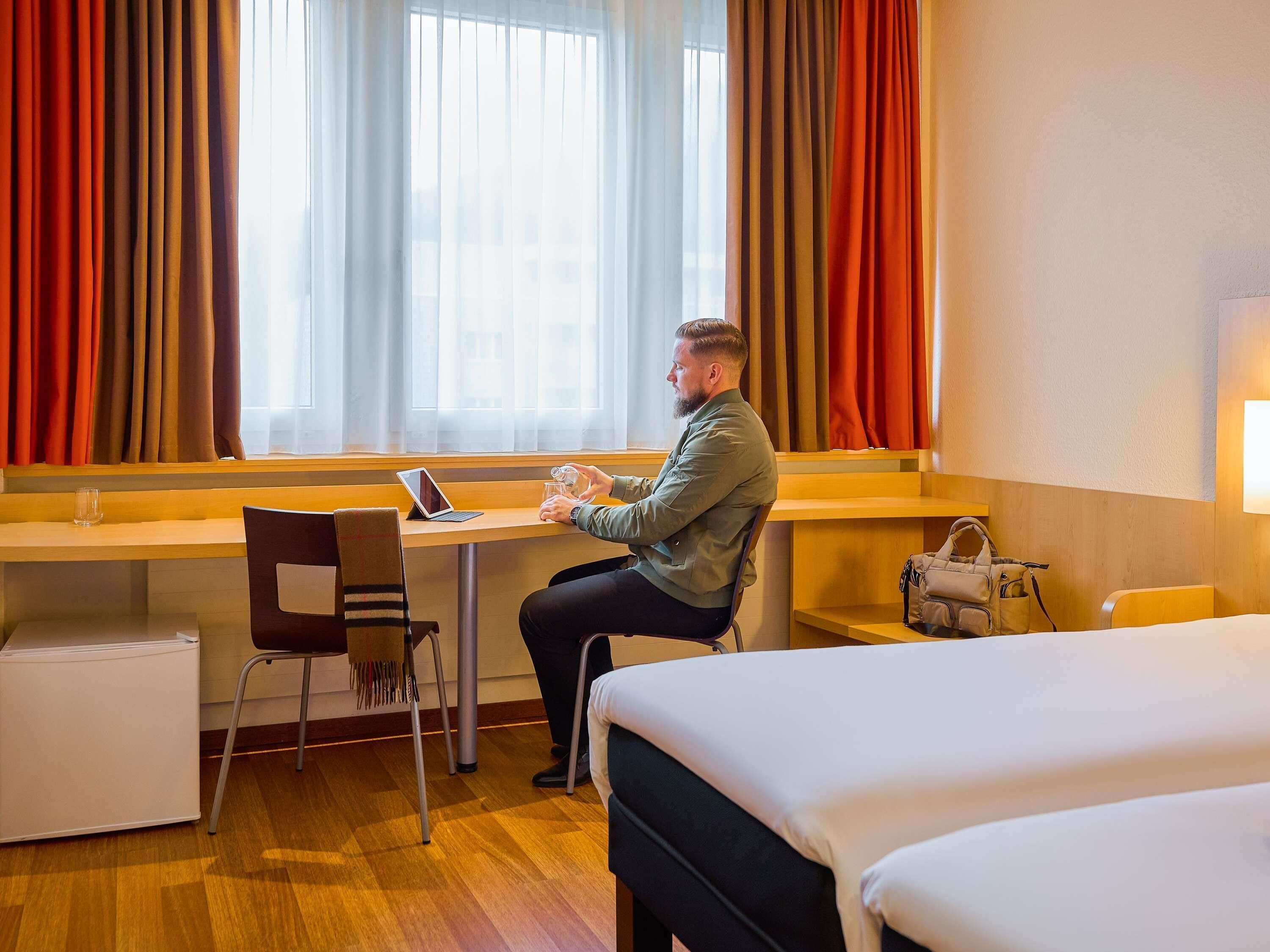 Habitación de hotel con cama, escritorio, computadora portátil, silla, una mesita y maleta.