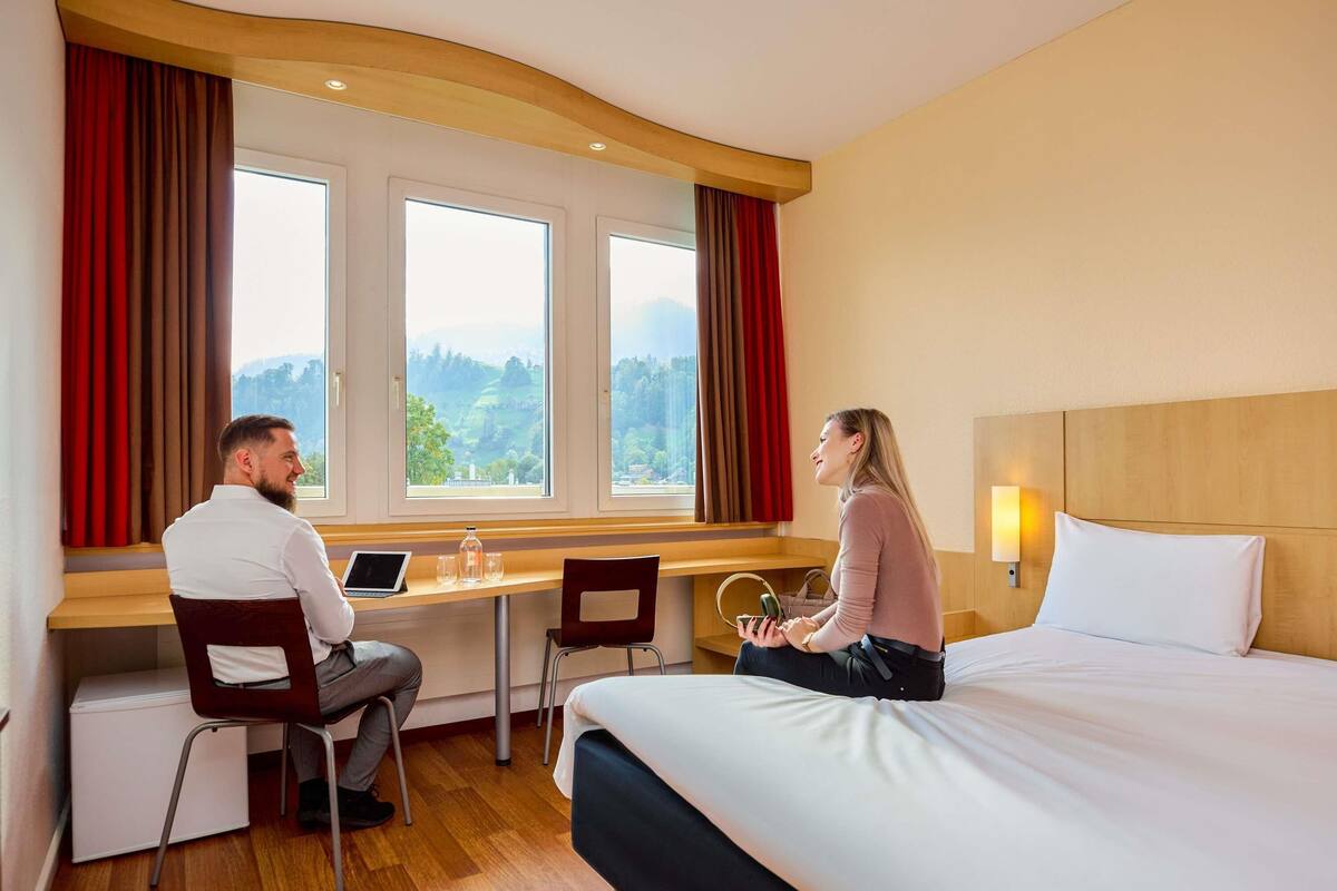 Une chambre d’hôtel avec un lit, un bureau et deux chaises. Il y a des fenêtres avec des rideaux, une vue sur l’extérieur et une lampe fixée au mur.