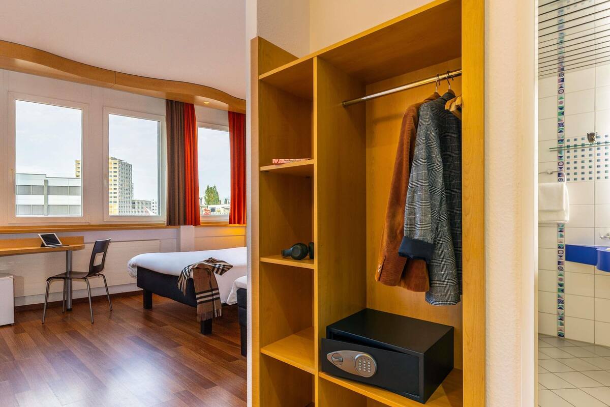 Une chambre d’hôtel avec un lit, un bureau, une armoire et une vue sur les bâtiments par la fenêtre.