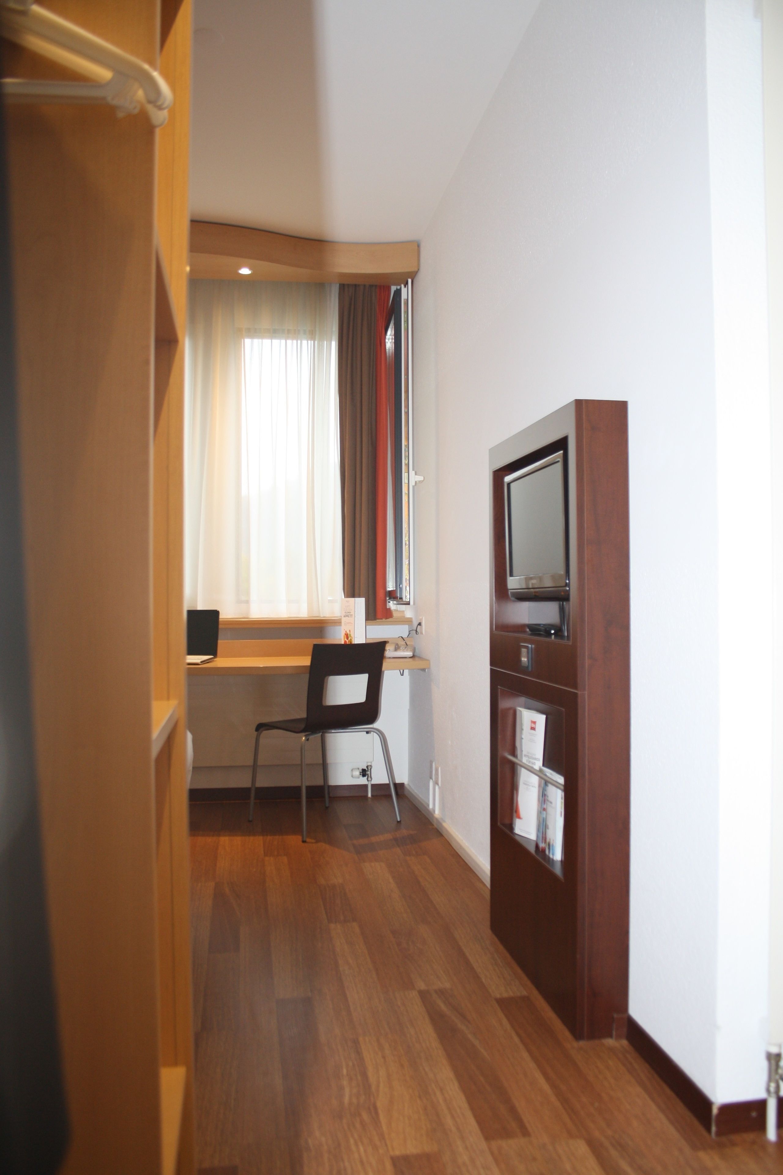 Una habitación con piso de madera, un escritorio con silla, un televisor y una ventana con cortinas.
