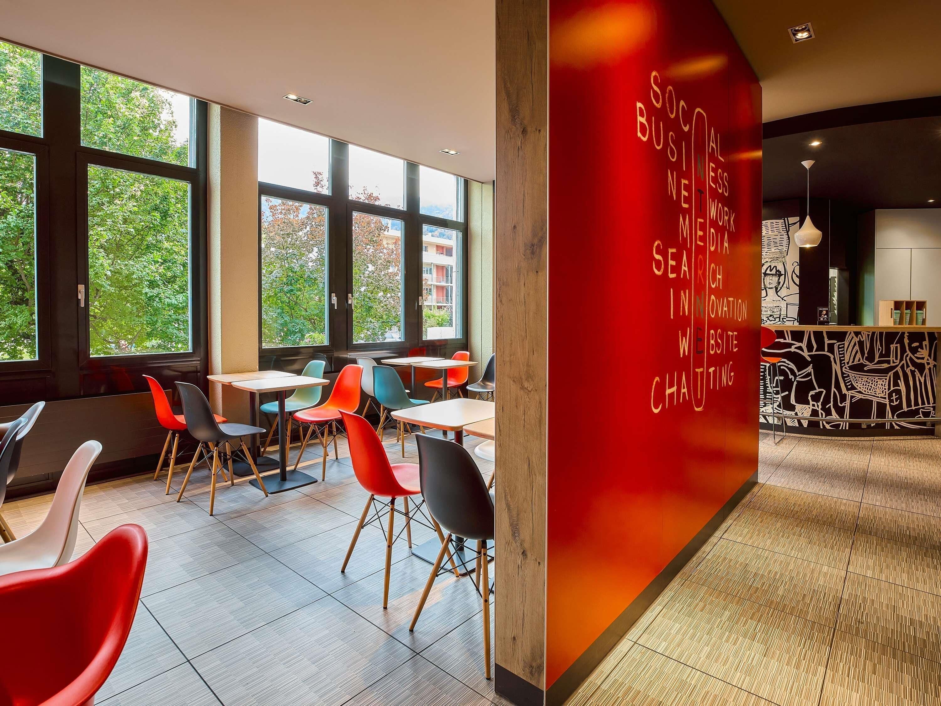 Una cafetería moderna con ventanas amplias, sillas coloridas y una pared roja con texto.