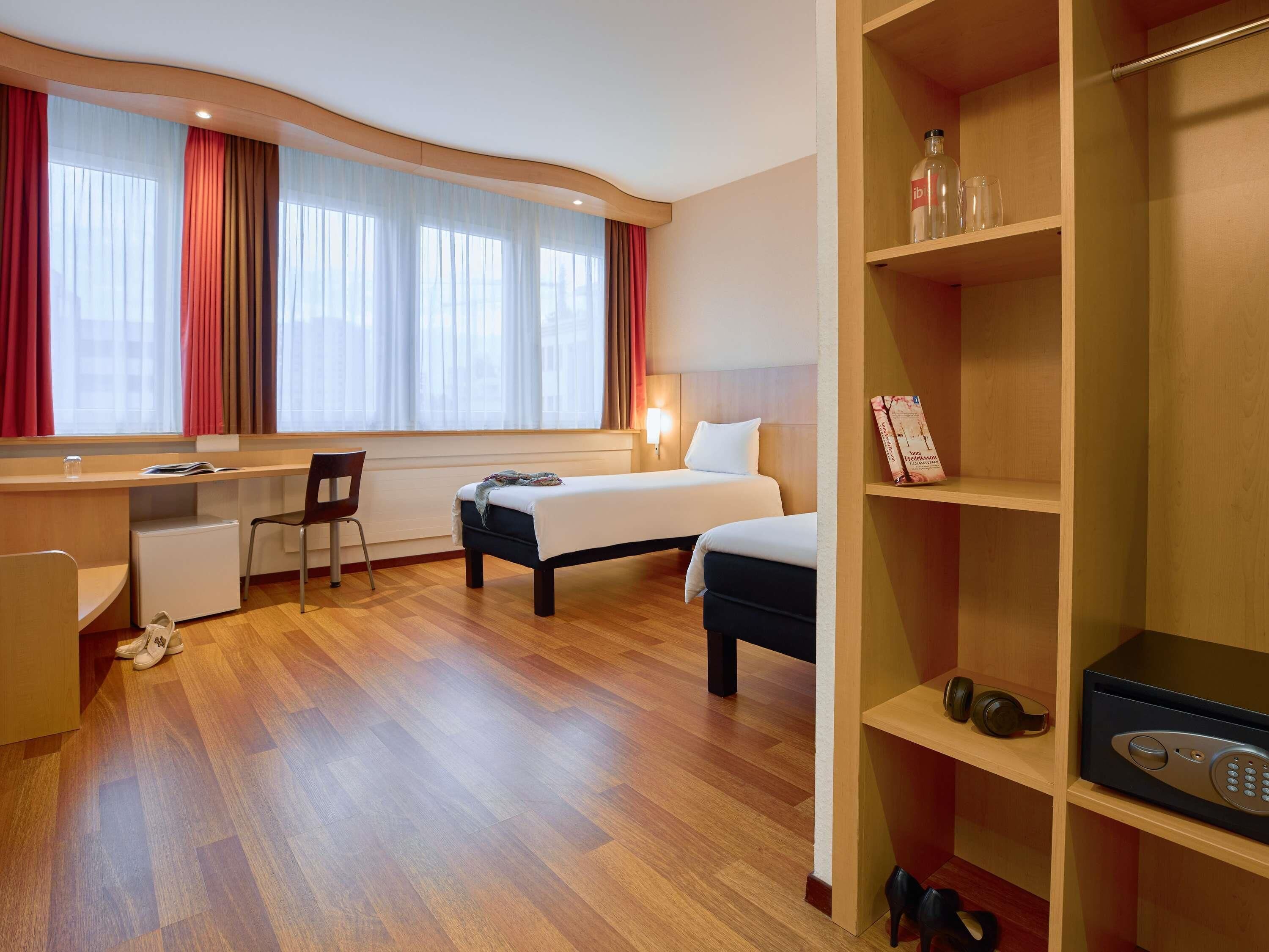 A hotel room with a bed, a desk, a chair, a small refrigerator, and a shelf with a remote control and headphones.