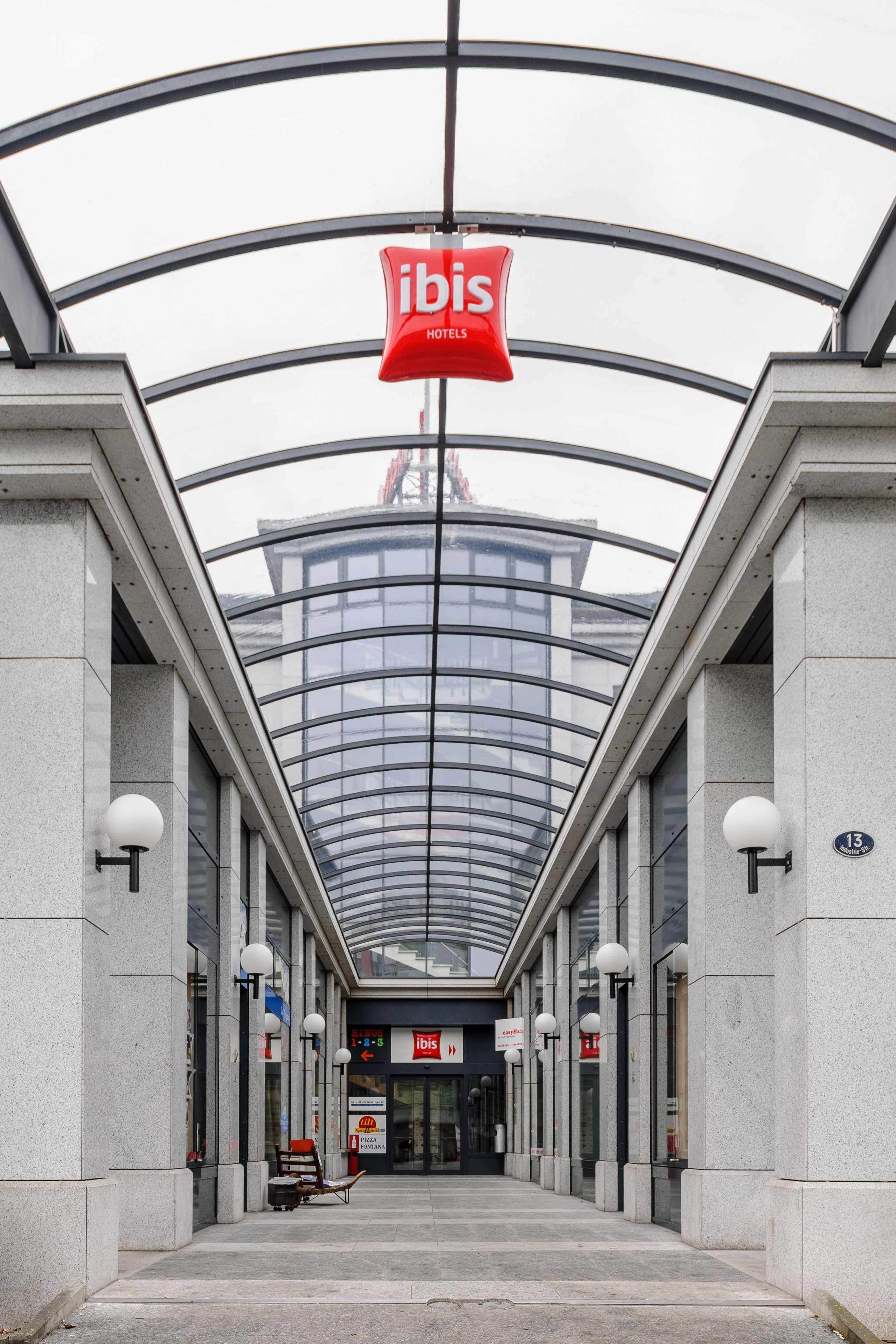 Ein moderner Hotelzugang mit Glaskuppel, einem roten 'ibis Hotels'-Schild und der Hausnummer 13 an der Wand.