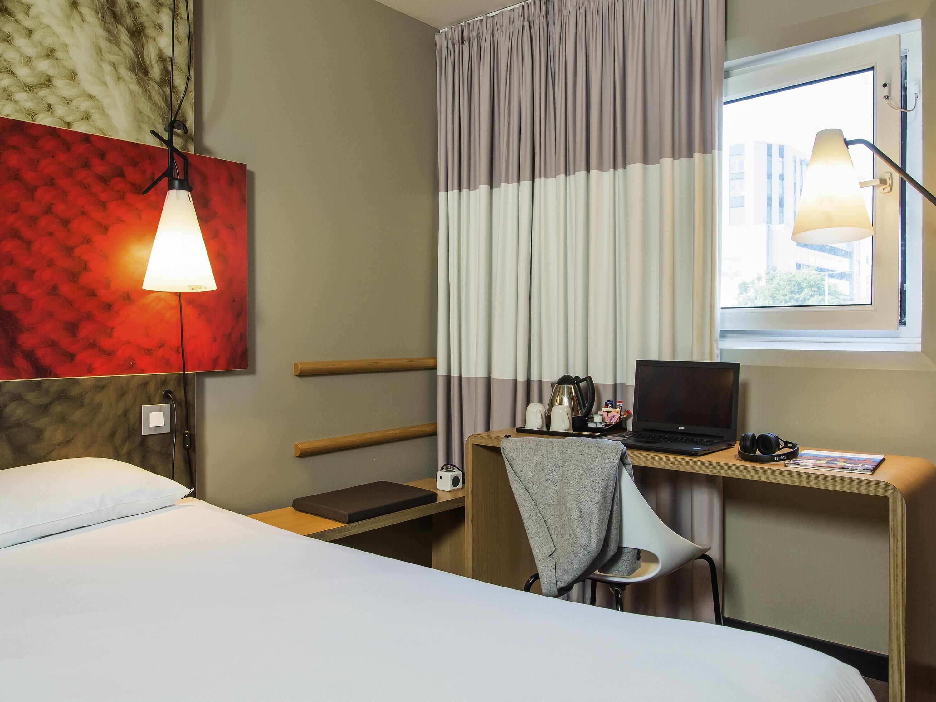 A hotel room with a bed, a desk with a laptop, a chair, a lamp, and a window with curtains.