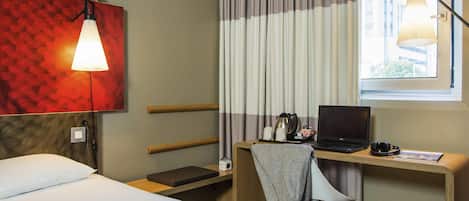 A hotel room with a bed, a desk with a laptop, a chair, a lamp, and a window with curtains.