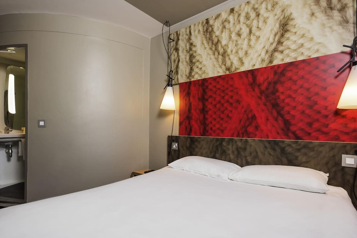Una habitación de hotel con cama, mesita de noche, lámpara de pared y espejo.