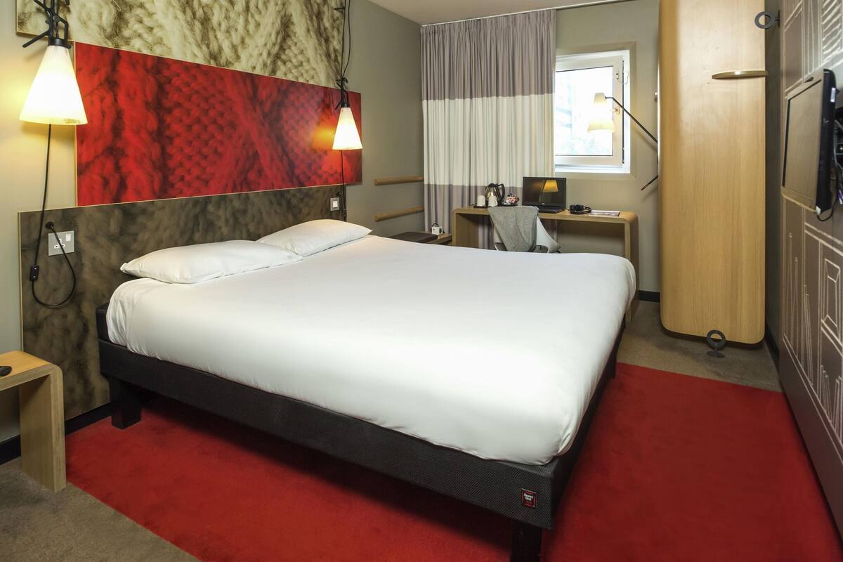 A hotel room with a large bed, a desk with a computer, a chair, a TV, and a wardrobe.