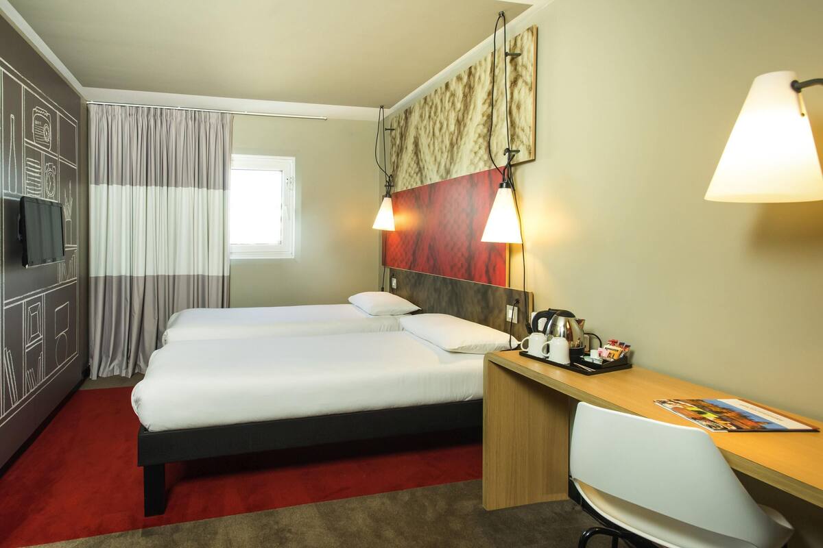 A hotel room with a double bed, a desk with a chair, a television, and a window with curtains.