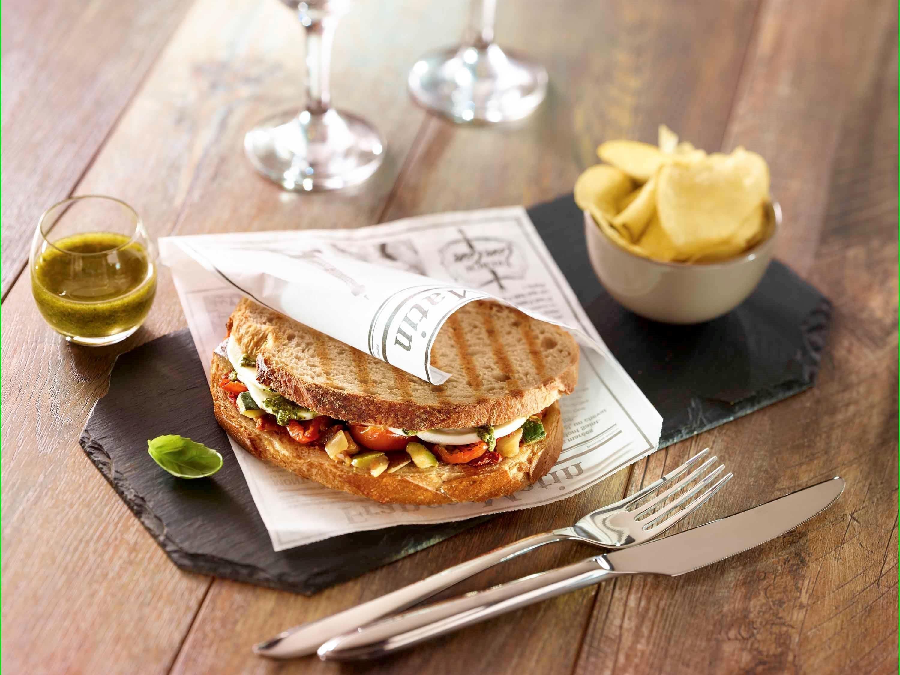 Un panino su un tagliere in ardesia, con una forchetta e un coltello accanto, un bicchiere di salsa verde e una ciotola di patatine.