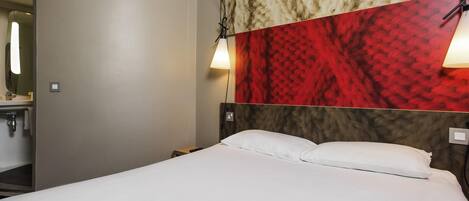 Una habitación de hotel con cama, mesita de noche, lámpara de pared y espejo.