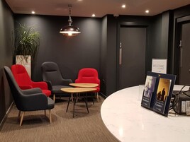 A modern hotel lobby with a reception desk, two red and one black armchairs, a small round table, a potted plant, and a 'Welcome' sign.