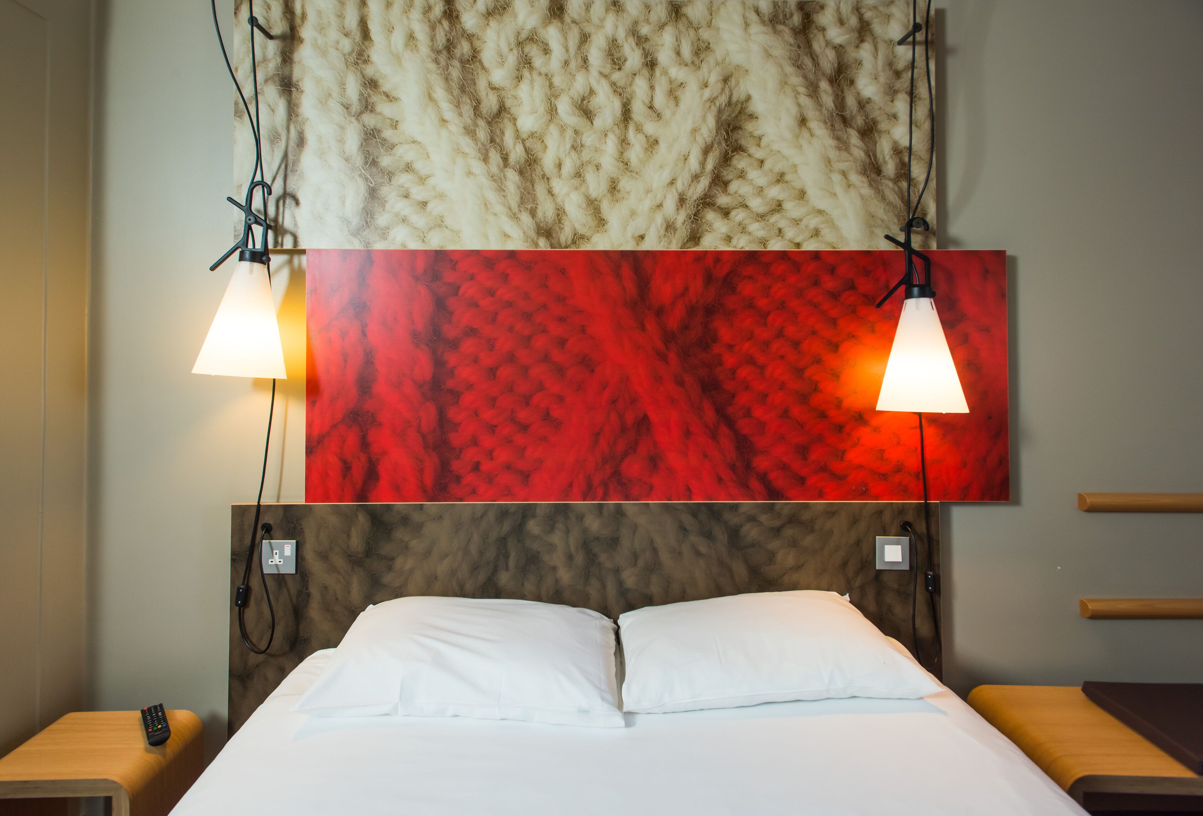 Una habitación de hotel con una cama, dos lámparas de noche, un control remoto sobre una mesita de noche y una pared texturizada en rojo y beige sobre la cama.
