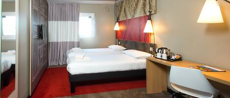 A hotel room with a double bed, a desk with a chair, a mirror, a wardrobe, and a red carpet.