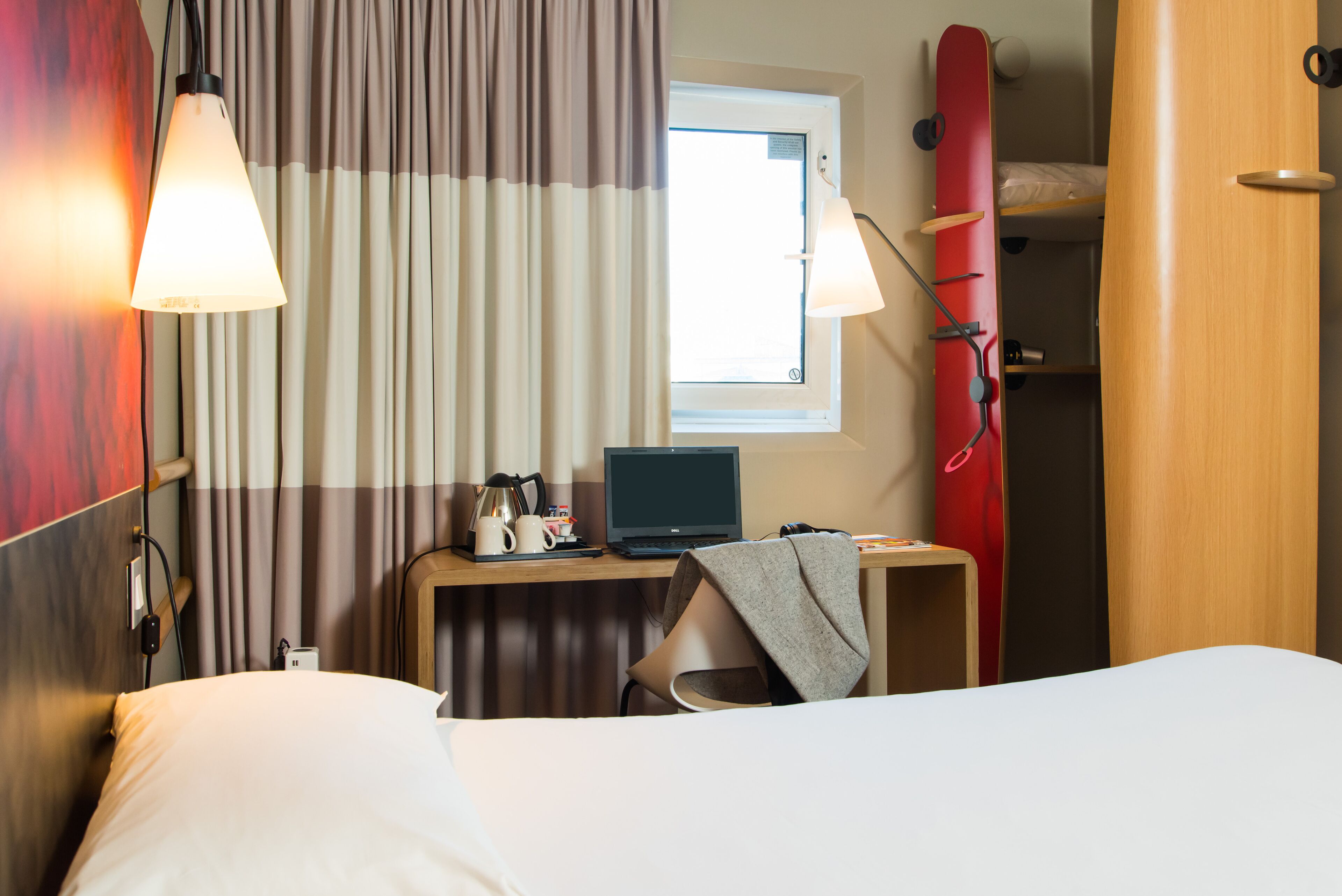 Habitación de hotel con cama, escritorio con una computadora portátil, una tabla de surf roja y una ventana con cortinas.