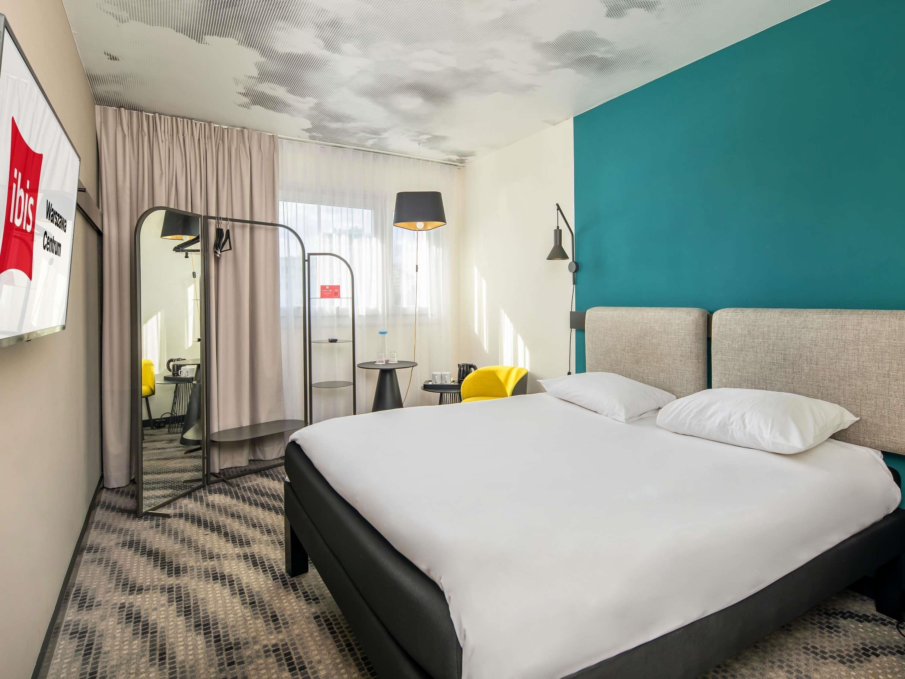 A hotel room with a bed, a desk, a chair, a lamp, and a view of the outside.