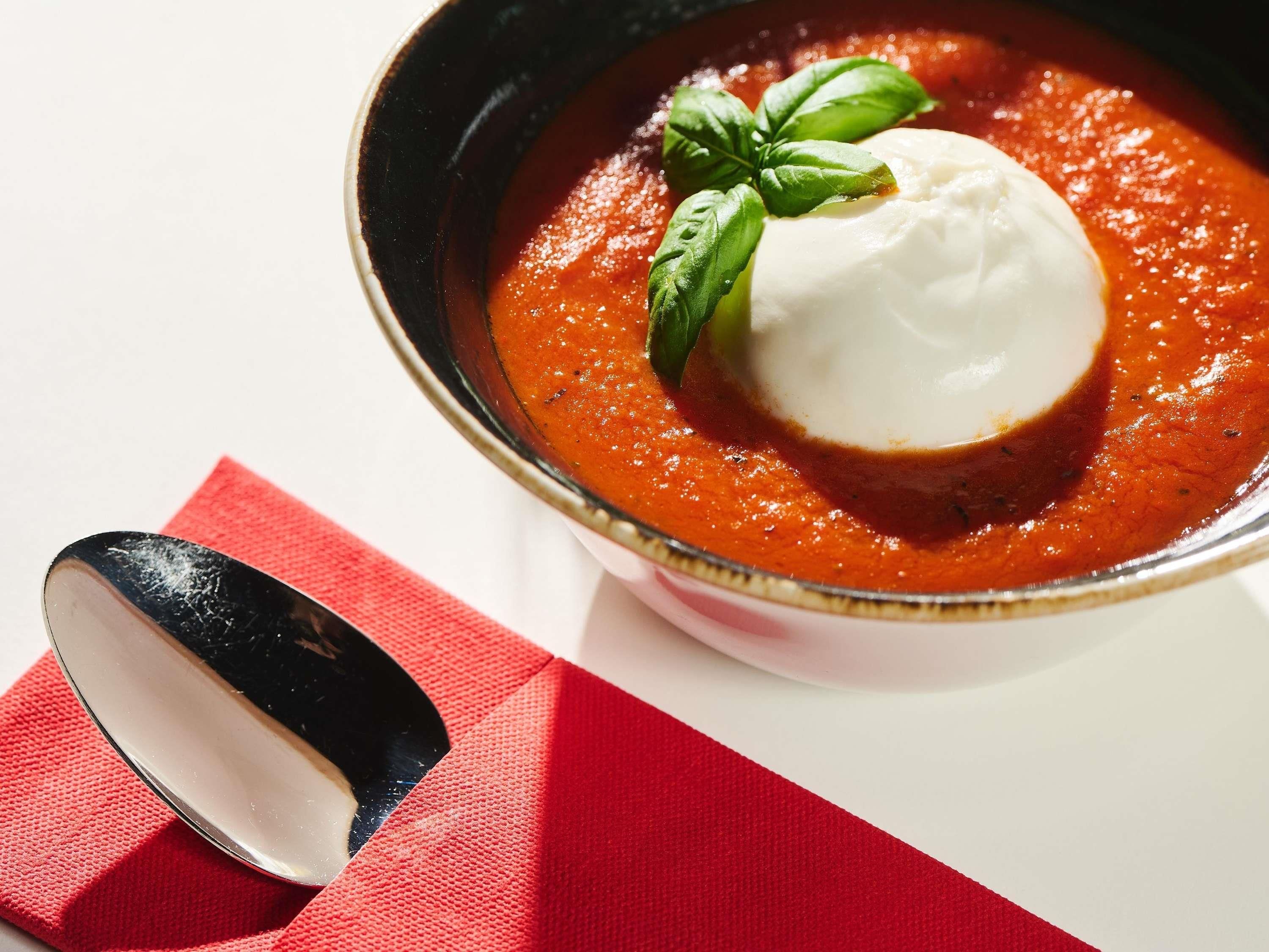 A bowl of tomato soup with a dollop of cream and a basil leaf on top, placed on a white surface with a red napkin beside it.