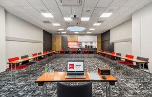 A conference room with a laptop displaying 'ibis Warszawa Centrum' on the screen, multiple tables with chairs, and a kitchen area in the background. - ibis Warszawa Centrum (Warsaw)