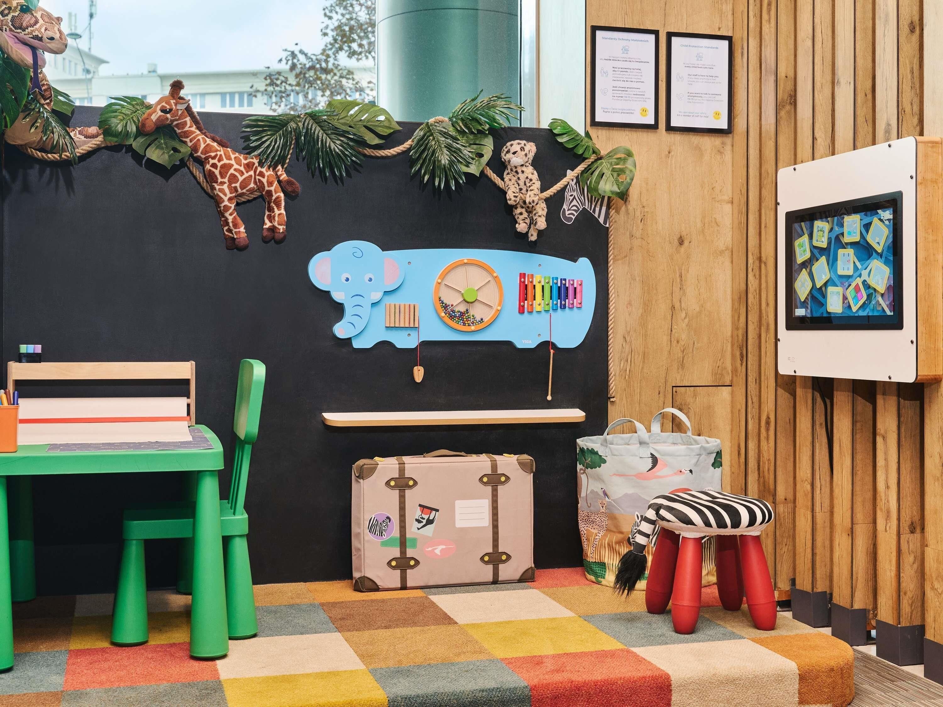 A children's play area with a black backdrop, a wooden desk, green chairs, a colorful rug, and a suitcase with travel stickers.