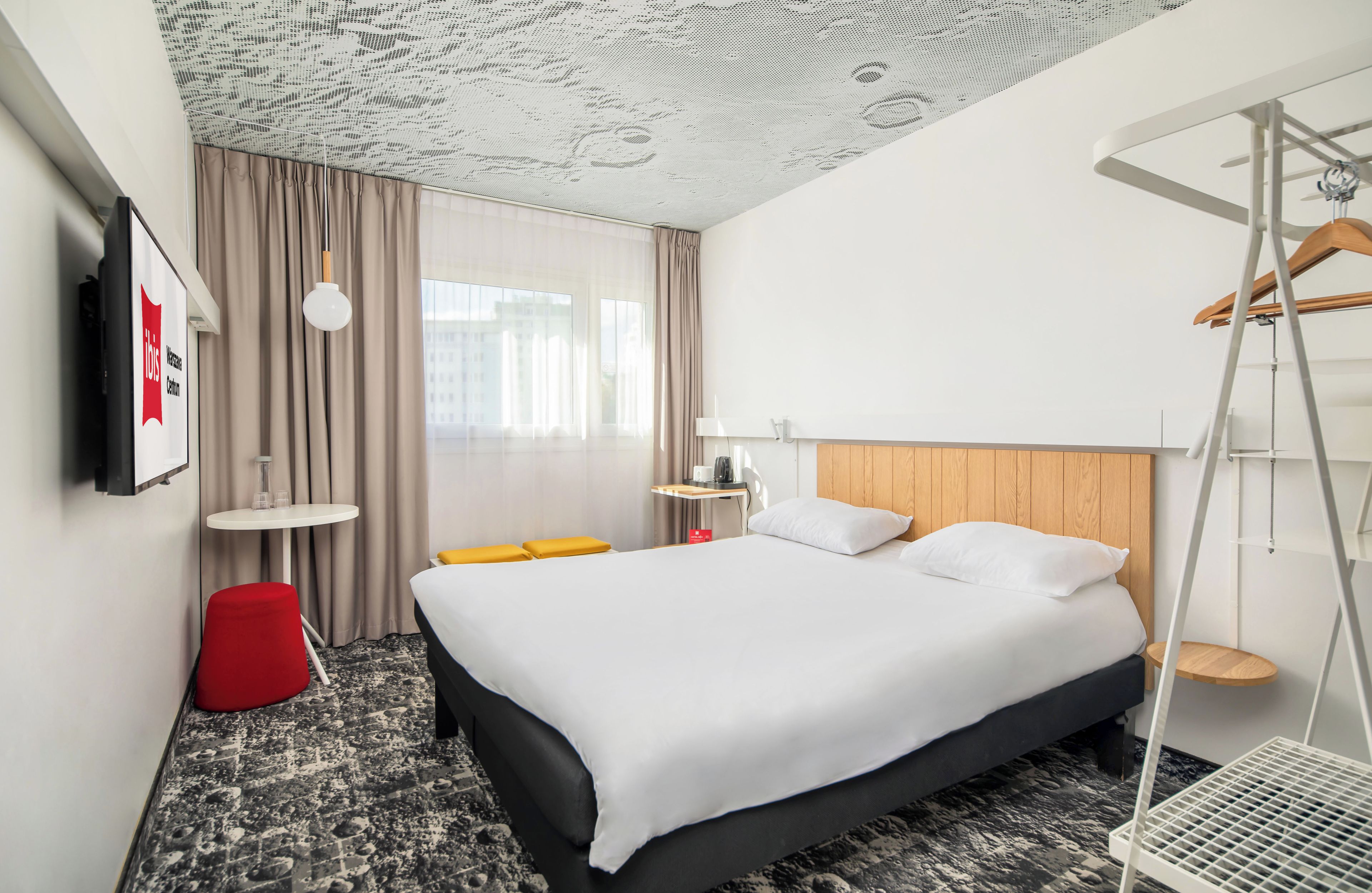 Habitación de hotel con cama, una mesita, una silla roja, una lámpara colgante y vistas a edificios a través del ventanal.