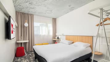 Habitación de hotel con cama, una mesita, una silla roja, una lámpara colgante y vistas a edificios a través del ventanal.