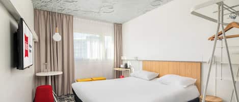 Habitación de hotel con cama, una mesita, una silla roja, una lámpara colgante y vistas a edificios a través del ventanal.
