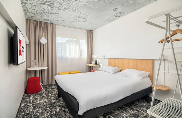 Habitación de hotel con cama, una mesita, una silla roja, una lámpara colgante y vistas a edificios a través del ventanal.