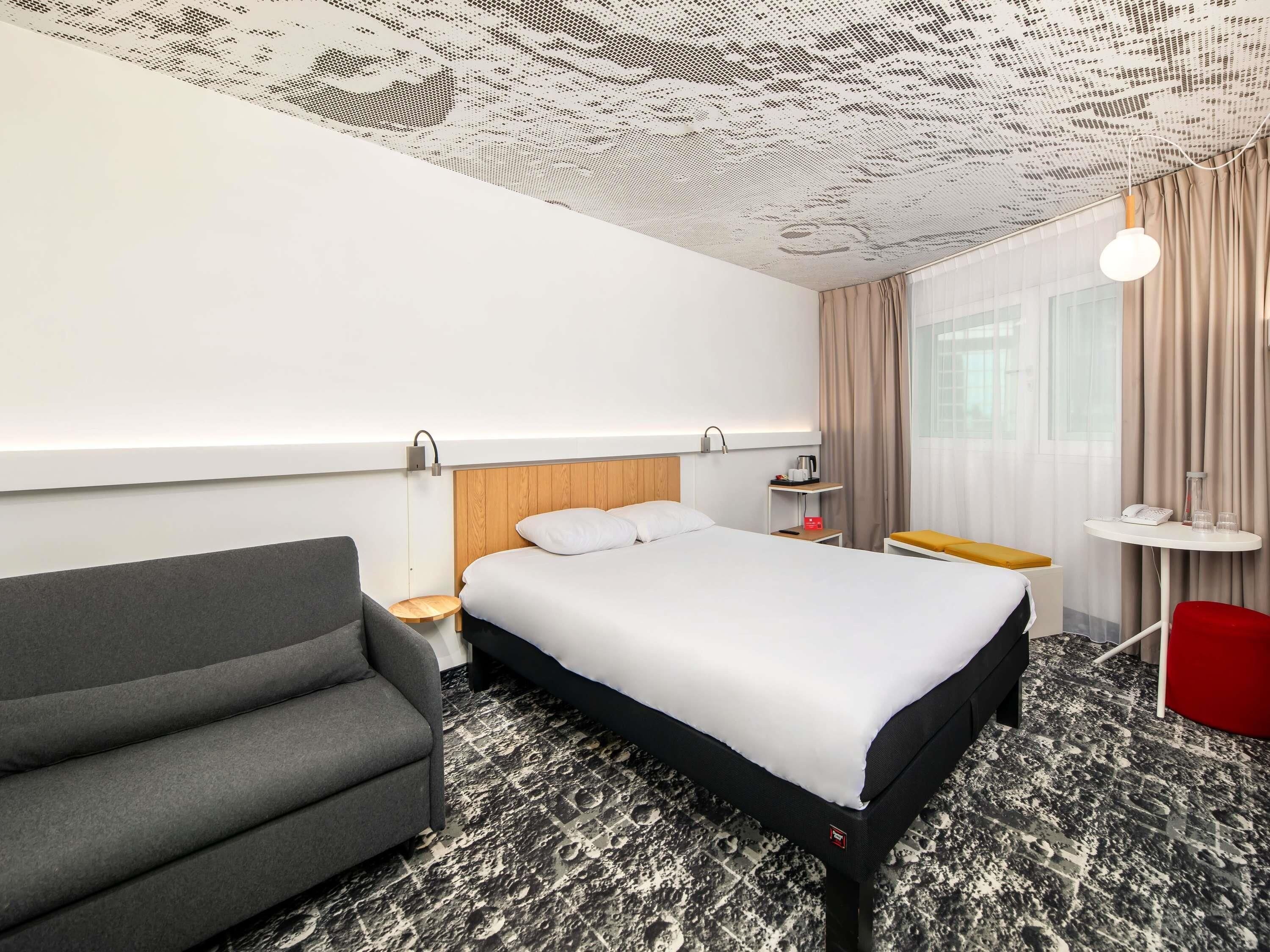 A hotel room with a bed, a grey armchair, a small table with a lamp, and a window with curtains.