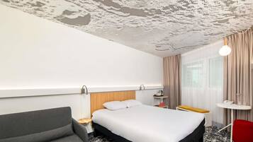 A hotel room with a bed, a grey armchair, a small table with a lamp, and a window with curtains.