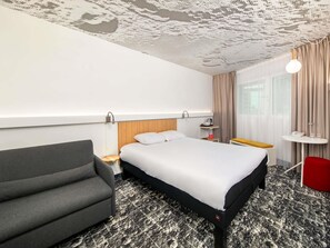 A hotel room with a bed, a grey armchair, a small table with a lamp, and a window with curtains. - ibis Warszawa Centrum (Warsaw)