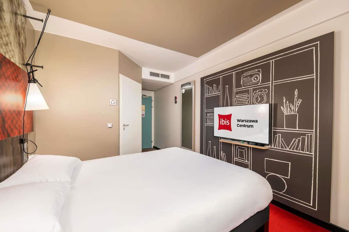 Habitación de hotel con una cama, un televisor montado en la pared y una alfombra roja.