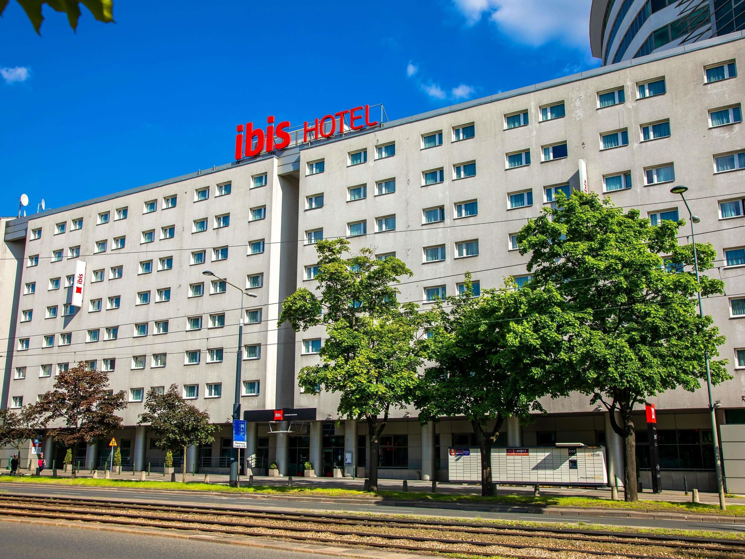 A modern hotel building with a prominent 'ibis HOTEL' sign, multiple windows, and a parking area with a train track in the foreground.