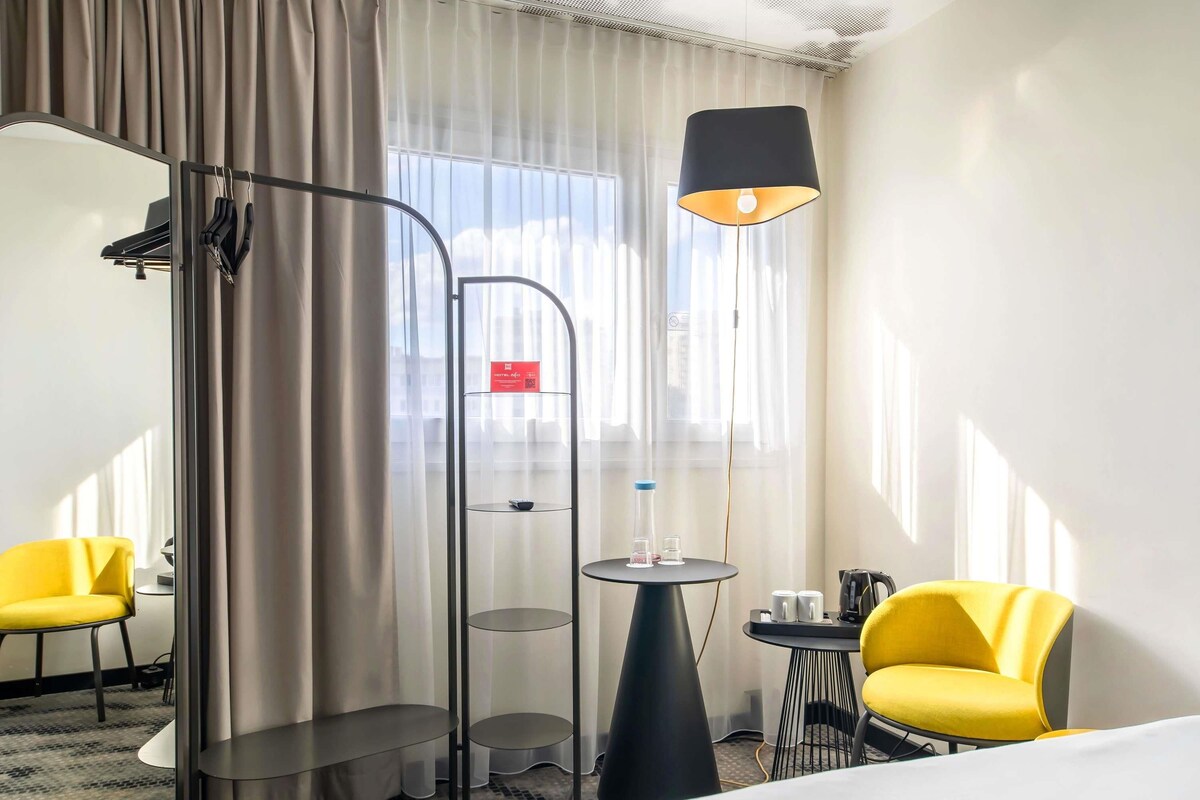 A hotel room with a bed, two yellow chairs, a small table with a lamp, and a glass shelf unit.