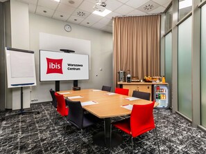A conference room with a large screen displaying 'ibis Warszawa Centrum,' a wooden table with chairs, a clock, a whiteboard, and a refrigerator with beverages. - ibis Warszawa Centrum (Warsaw)