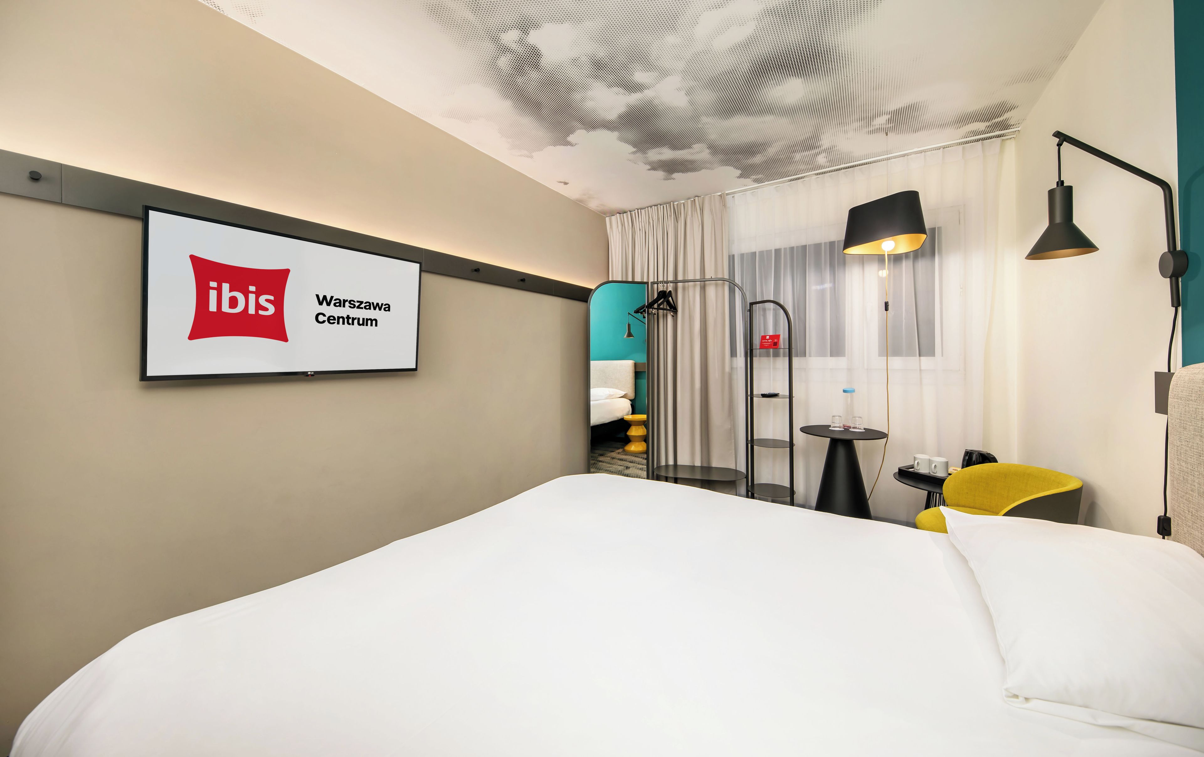 A hotel room with a bed, a desk, a chair, a lamp, a TV screen displaying the ibis logo, and a ceiling with a cloud design.