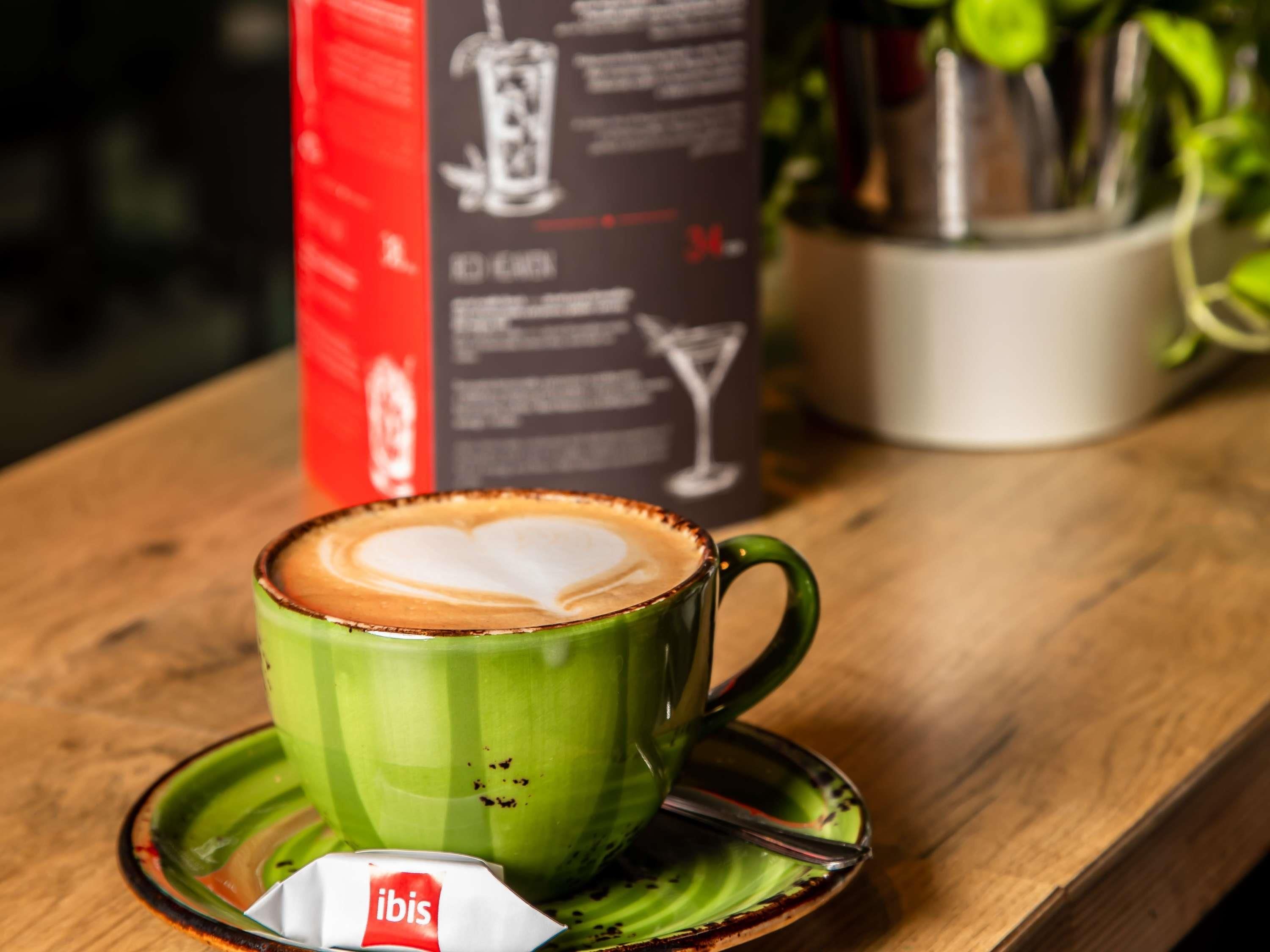 A green cup of coffee with a heart-shaped latte art on a saucer, a small packet of ibis tea, and a menu stand in the background.