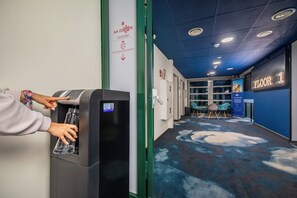 A water dispenser with a hand filling a bottle. A sign on the wall reads 'NA ZDROWIE' and 'TOGETHER WE CAN MAKE POSITIVE CHANGES!' The hallway has a blue carpet and a sign indicating 'FLOOR 1'. - ibis Warszawa Centrum (Warsaw)