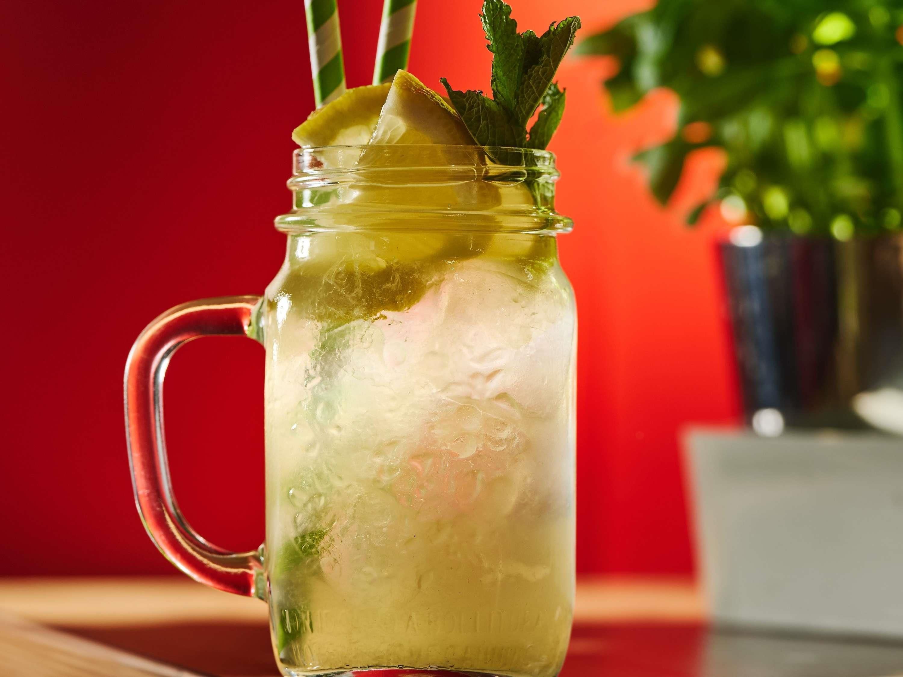 Un vaso de vidrio transparente con asa, lleno de una bebida y hielo, adornado con una rodaja de lima y hojas de menta, sobre un fondo rojo.