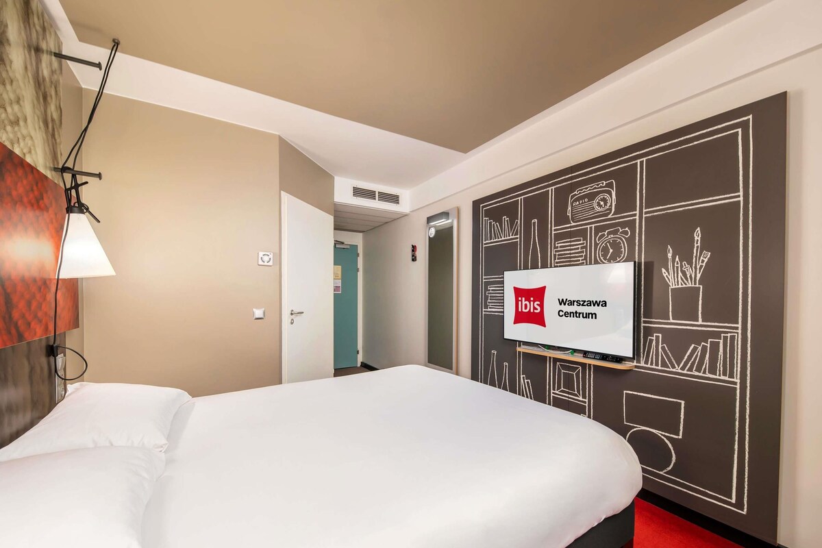 A hotel room with a bed, a wall-mounted TV, and a red carpet.
